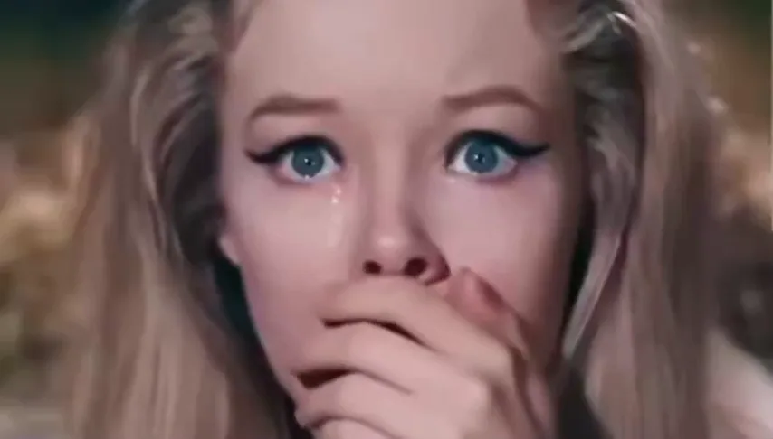 Blonde woman with wide blue eyes covering her mouth in surprise as she watches