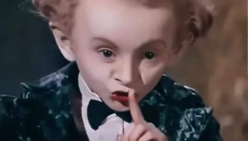 Curly-haired performer in a dark sequined tailcoat and bow tie raising a finger to his lips in a hay-filled barn